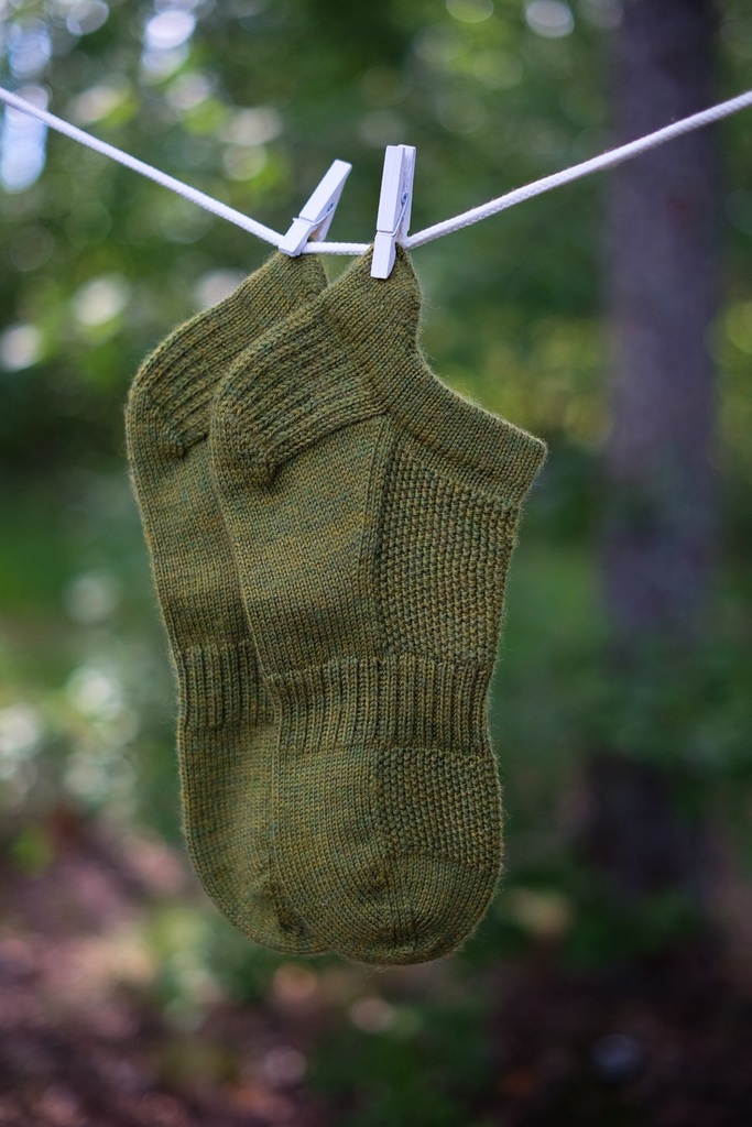 Green So Sporty Socks hanging on a clothesline with clothespins, surrounded by green foliage in the background.